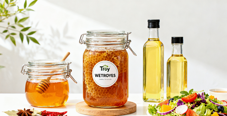 A stunning hero image showing a diverse array of Troy's glass jars and bottles filled with various food products like artisanal honey, vibrant spices, colorful salads, and specialty oils. The composition should be bright, clean, and appealing